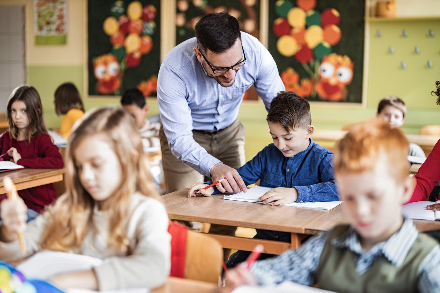 Ein Grundschullehrer steht einem Schülertisch im Klassenzimmer. Er beugt sich zu einem Jungen und zeigt auf eine Aufgabe im Heft. Im Klassenzimmer vor und hinter dem Jungen sitzen Kinder an ihren Schülertischen, die selbstständig die Aufgabenstellung bearbeiten.